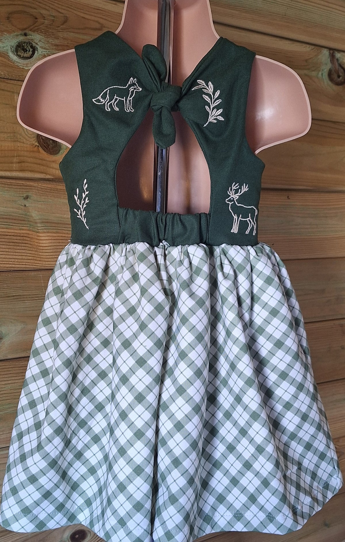 A bow back baby and toddler dress with forest green 100% cotton bodice scattered with cream embroidered motifs. Motifs include fox, deer and leaves. The gathered skirt is a organic cotton green plaid fabric.