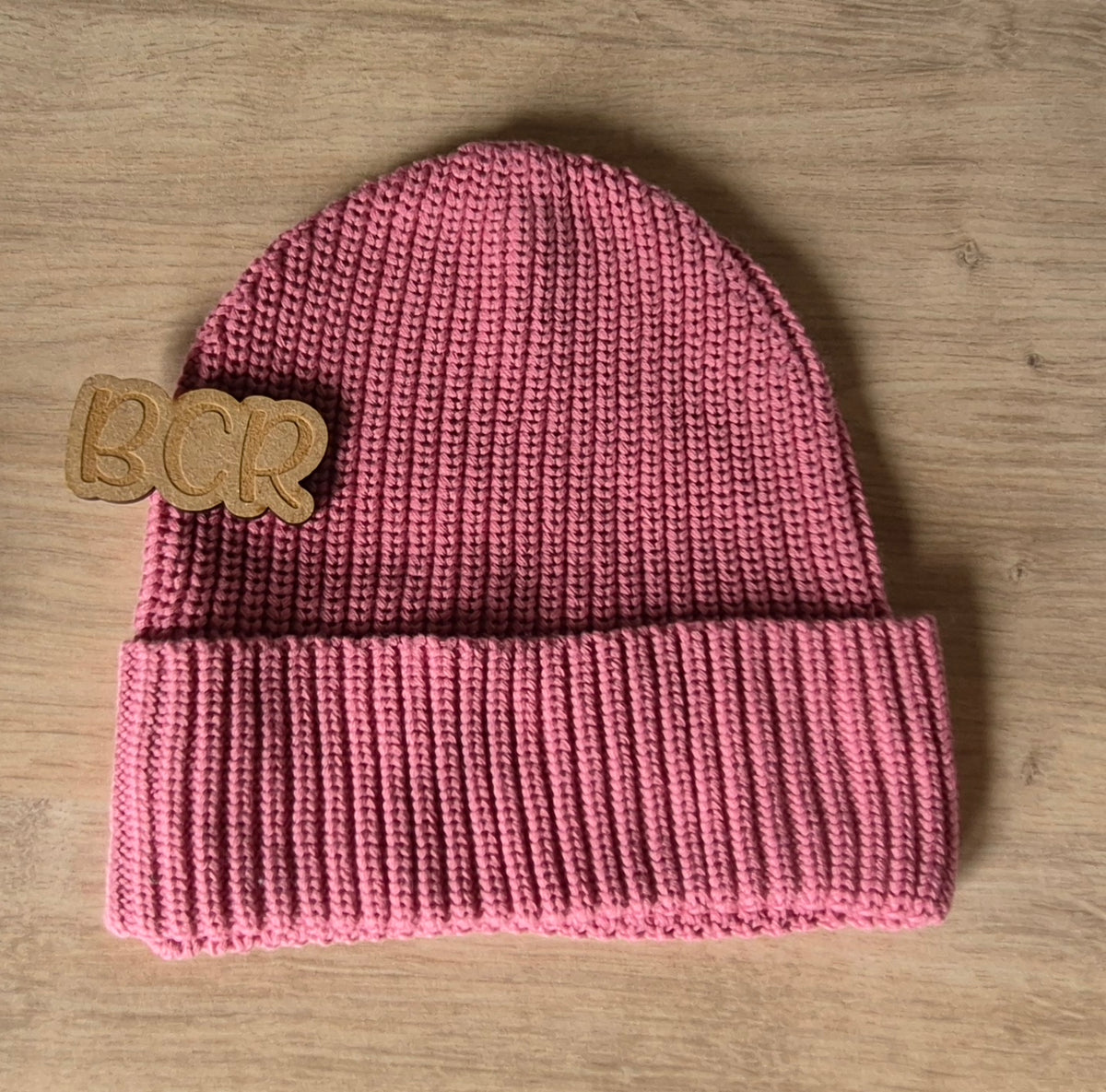 A pink ribbed knit beanie hat with a wooden 'BCR' logo patch on the side, displayed flat on a light wood grain surface.