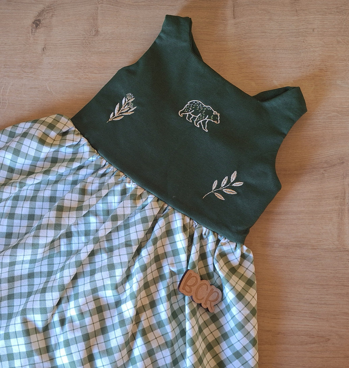 A bow back baby and toddler dress with forest green 100% cotton bodice scattered with cream embroidered motifs. Motifs include bear and leaves. The gathered skirt is organic cotton green plaid fabric. 