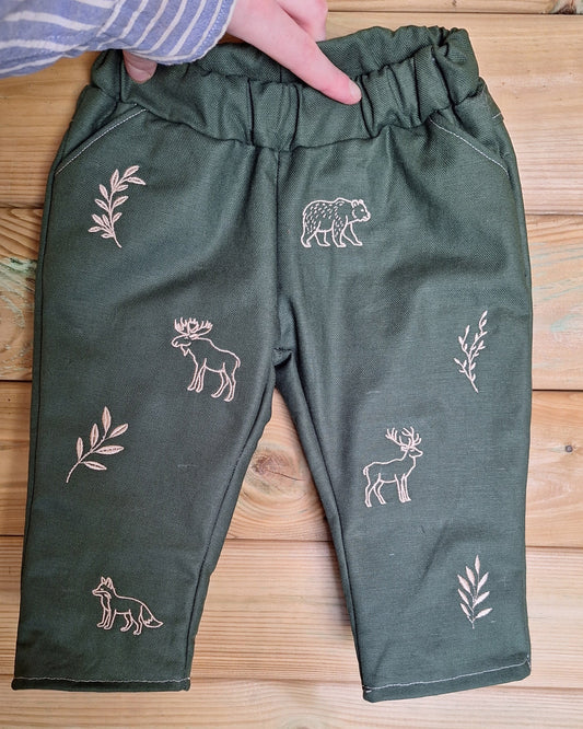 A pair of dark olive green children's pants with embroidered brown animal and leaf motifs, held by a hand over a light wood background.