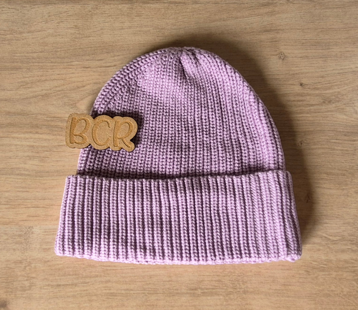 A light purple knit beanie hat with a folded cuff, featuring a wooden 'BCR' patch on the side, displayed flat on a light wood background.