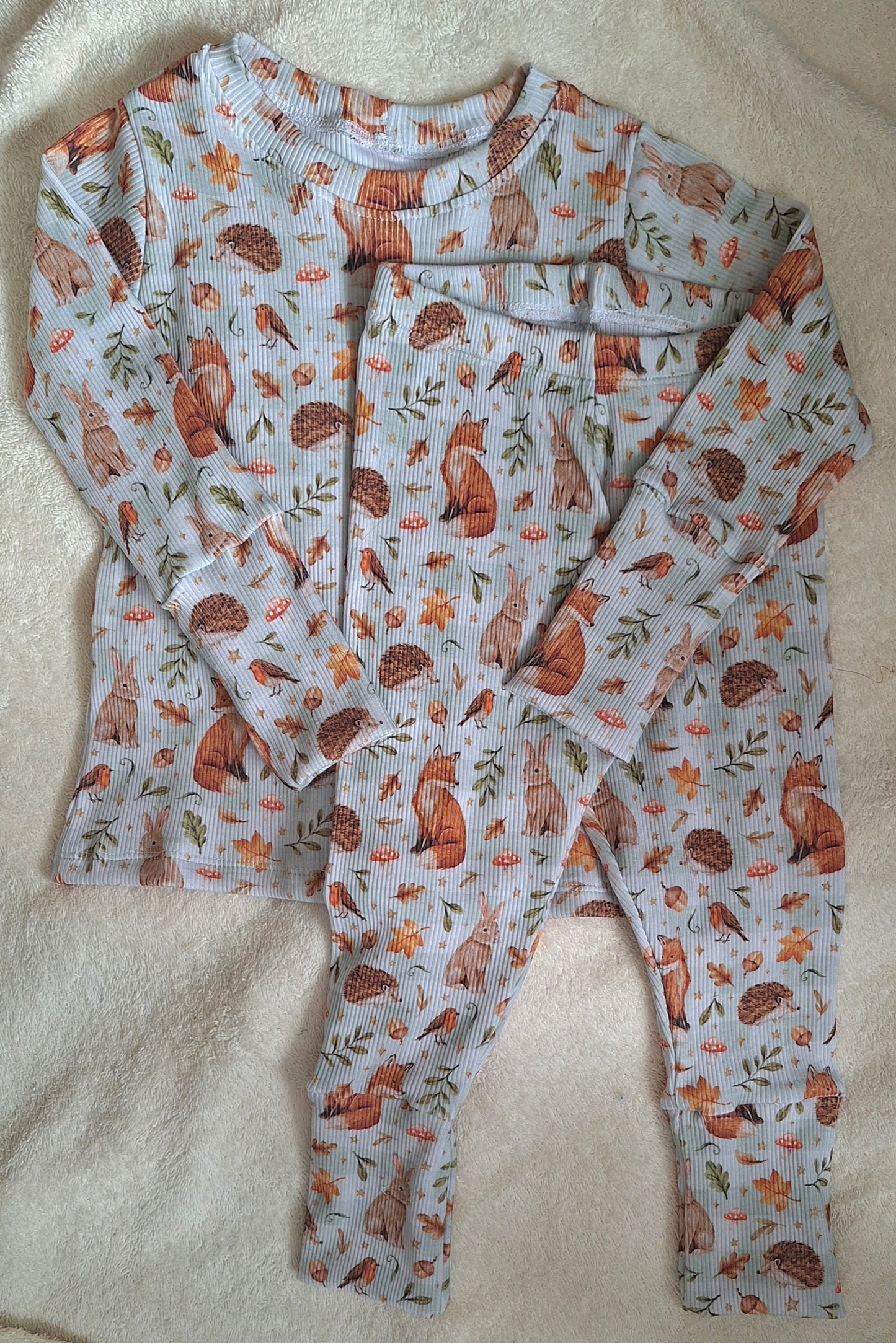A two-piece children's pajama set featuring a woodland print of foxes, rabbits, robins and hedgehogs, pine cones, and acorns on a light blue-gray background, laid flat on a white textured blanket.