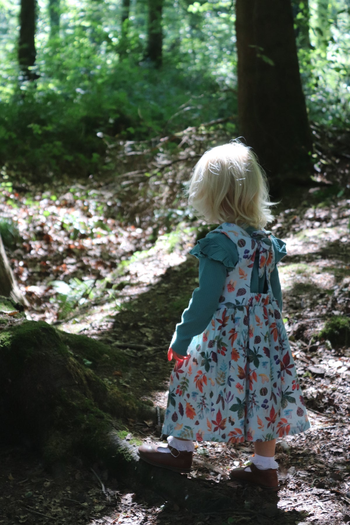 A young child with blonde hair seen from behind, wearing a floral print dress over a teal long-sleeve shirt, standing on a forest path.