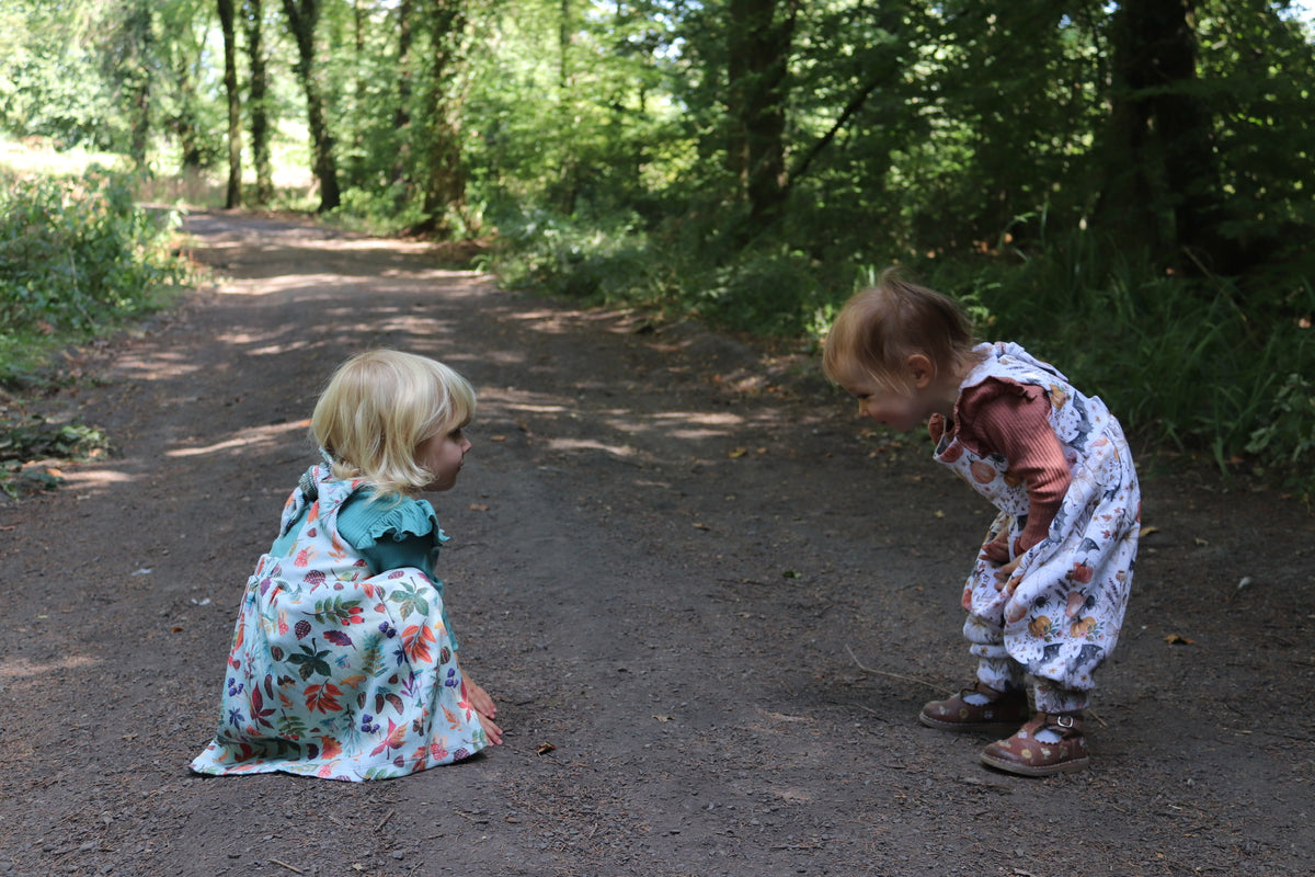 Two toddlers wearing a bow back dress with autumn leaves and a pretty Halloween bow back Napa romper  sit on a dirt path in a wooded outdoor setting.