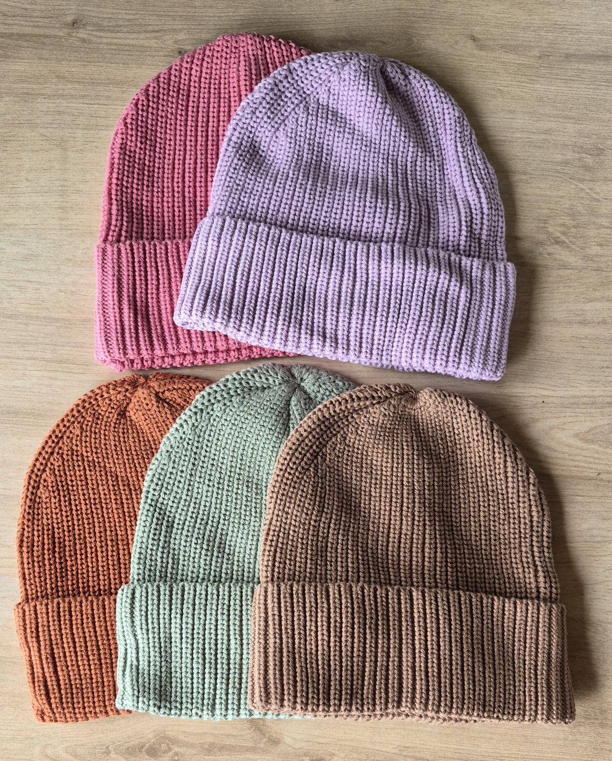 Five organic cotton ribbed knit beanies arranged in a fan shape on a light wood background. The top pair is pink and lavender, and the bottom pair is terracotta, sage green, and taupe.