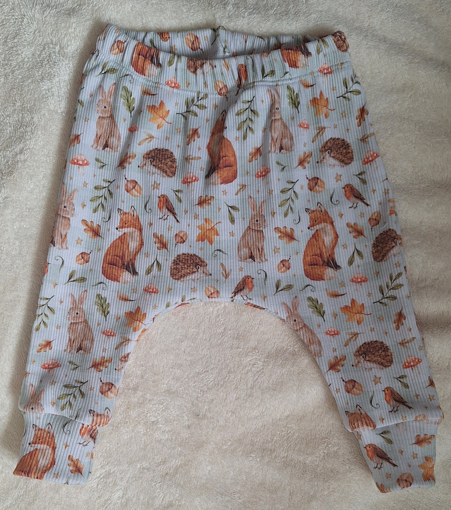 sage ribbed baby pants with an all-over woodland print of foxes, rabbits, hedgehogs, leaves, and mushrooms, laid flat on a beige textured blanket.
