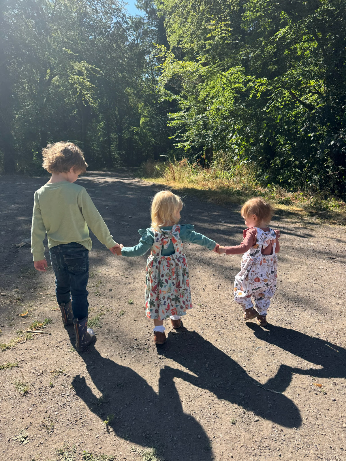 Three children holding hands walking away on a dirt path surrounded by green trees in bright sunlight.