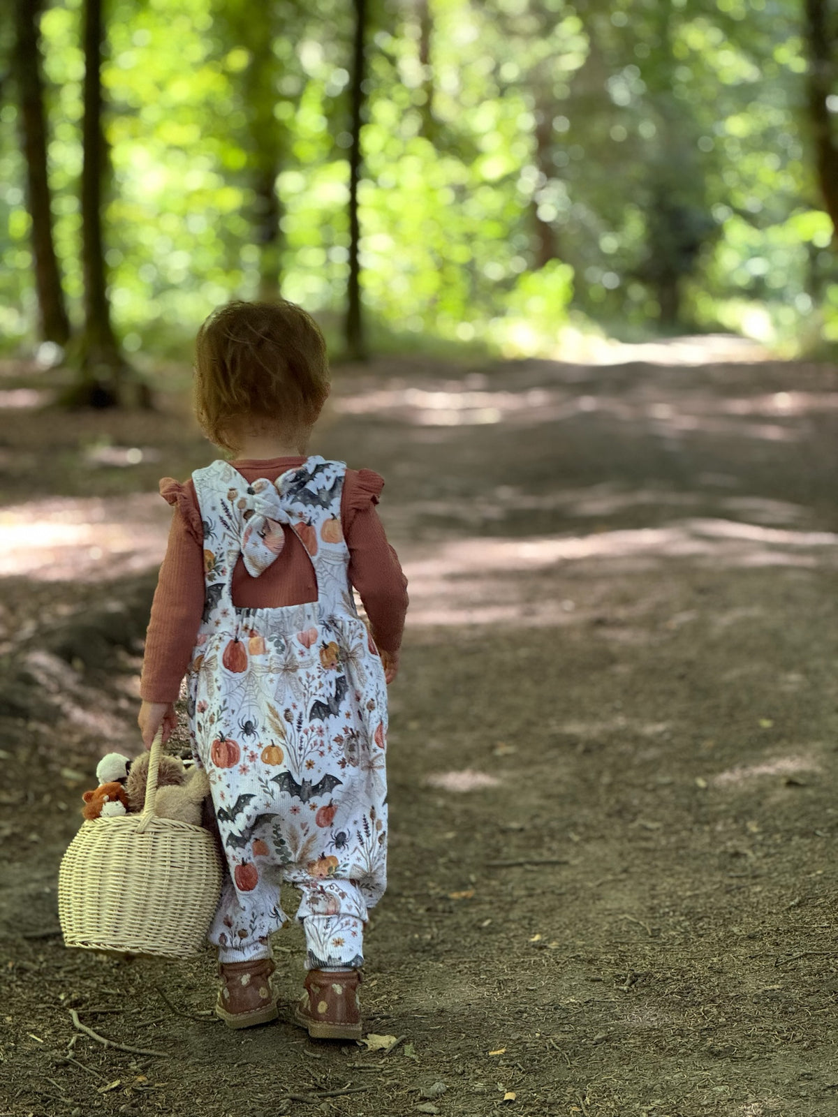 A toddler seen from behind wearing a white bow back Napa romper with a colourful pumpkin, Halloween and leaf print, carrying a small woven basket on a dirt path in a sunlit forest.