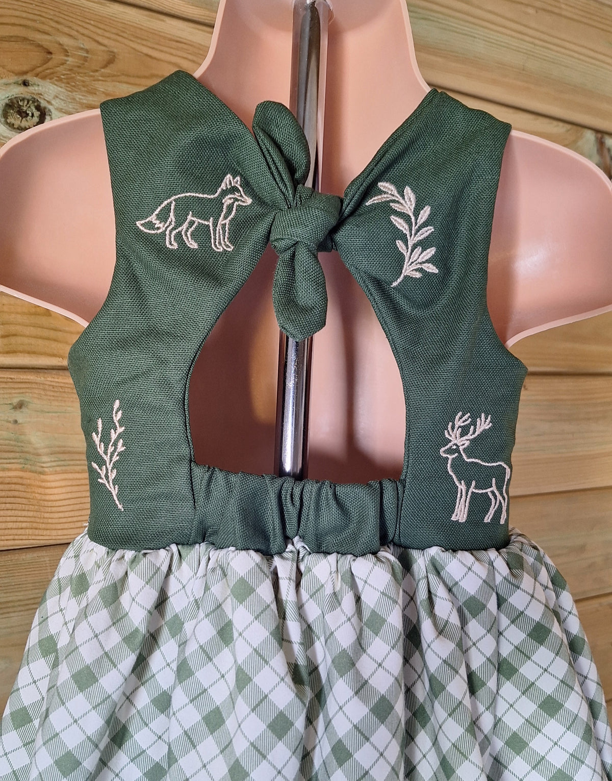 A close up of the back of the bow back baby and toddler dress with forest green 100% cotton bodice scattered with cream embroidered motifs. Motifs include fox, deer and leaves. The gathered skirt is a organic cotton green plaid fabric.