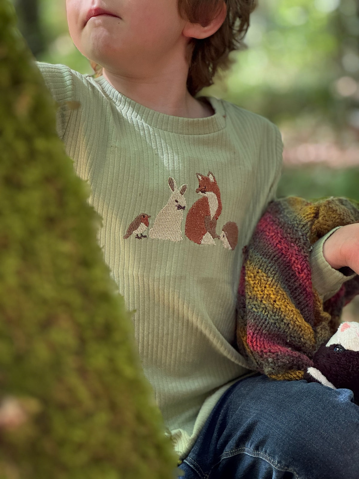 A child wearing a ribbed sage green long-sleeve top with a knitted woodland animal motif on the chest, holding a colorful striped knit blanket in an outdoor forest setting.