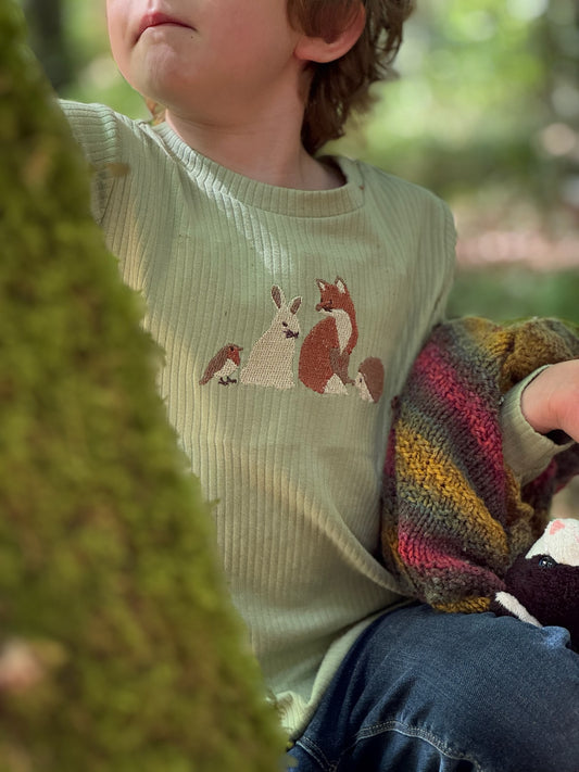 A child wearing a ribbed sage green long-sleeve top with a knitted woodland animal motif on the chest, holding a colorful striped knit blanket in an outdoor forest setting.