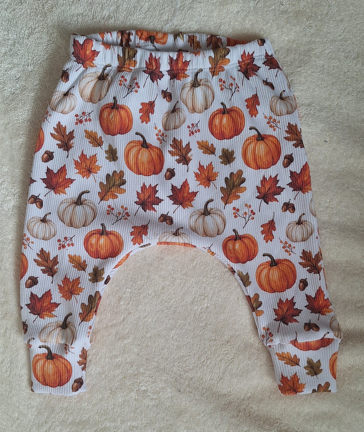 White ribbed baby pants with an all-over autumn print of pumpkins, maple leaves, acorns, and leaves in orange, brown, and beige, laid flat on a beige textured fabric background.